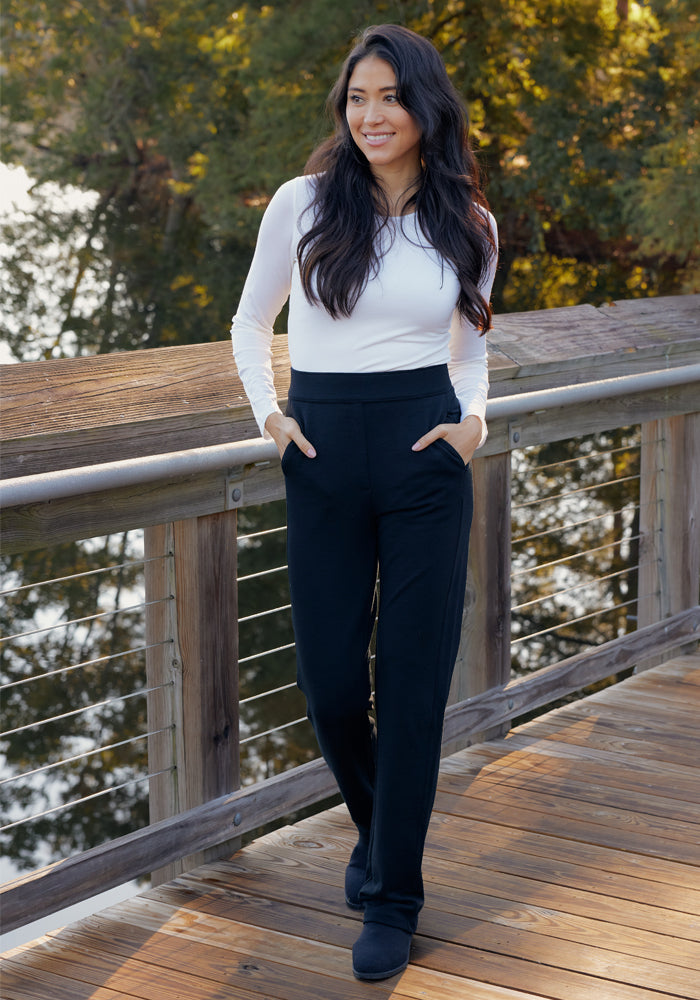 A woman with long dark hair, in a white long-sleeve top and Woolx Emerson Straight Leg Pants - Black, stands on a wooden bridge with her hands in her pockets. Trees and water are visible behind her.