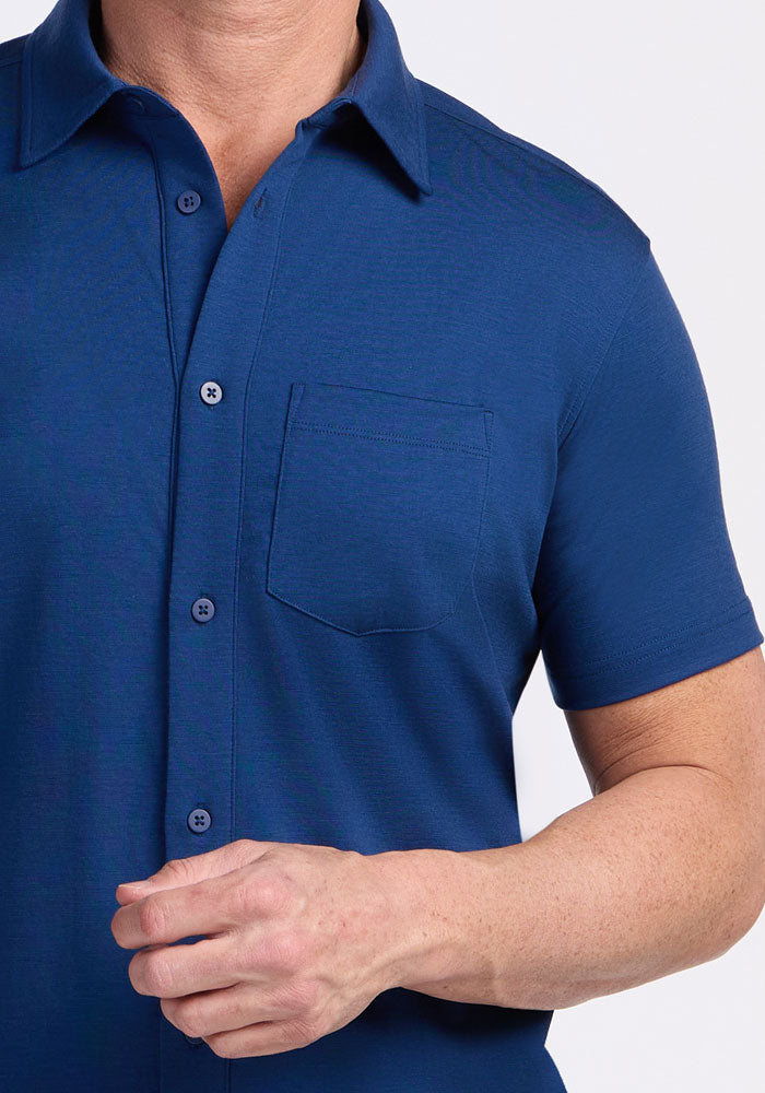 A person models the Woolx Endi Full Button Down Shirt in Estate Blue, featuring short sleeves and a chest pocket, against a plain white background. Only the upper torso and one hand are shown.