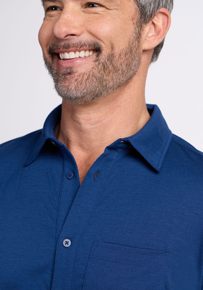 A smiling man with short gray hair and a beard models the Woolx Endi Full Button Down Shirt in Estate Blue, featuring a front pocket, shown from the shoulders up against a plain background.