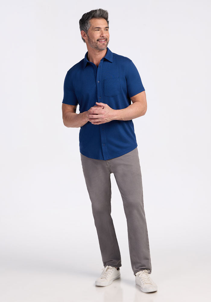 A man with short gray hair stands smiling against a plain white background, wearing gray pants, white sneakers, and the Woolx Endi Full Button Down Shirt in Estate Blue.