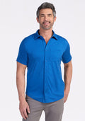 A smiling man with short gray hair and a beard wears the Woolx Endi Full Button Down Shirt in Glacial Blue and gray pants, standing against a plain white background with one hand in his pocket. | Matthew is 6’, wearing a size L