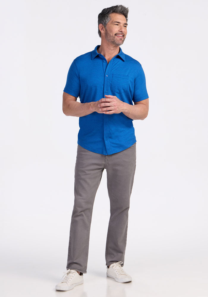 A man stands smiling with his hands clasped, wearing the Woolx Endi Full Button Down Shirt in Glacial Blue, paired with gray pants and white sneakers, against a plain white background.