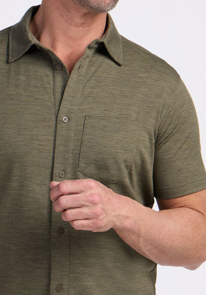 A man wearing the Woolx Endi Full Button Down Shirt in Sage Olive Melange, featuring short sleeves and a chest pocket, is shown from shoulders to mid-torso with his head partially out of frame as he holds the shirt near his chest.