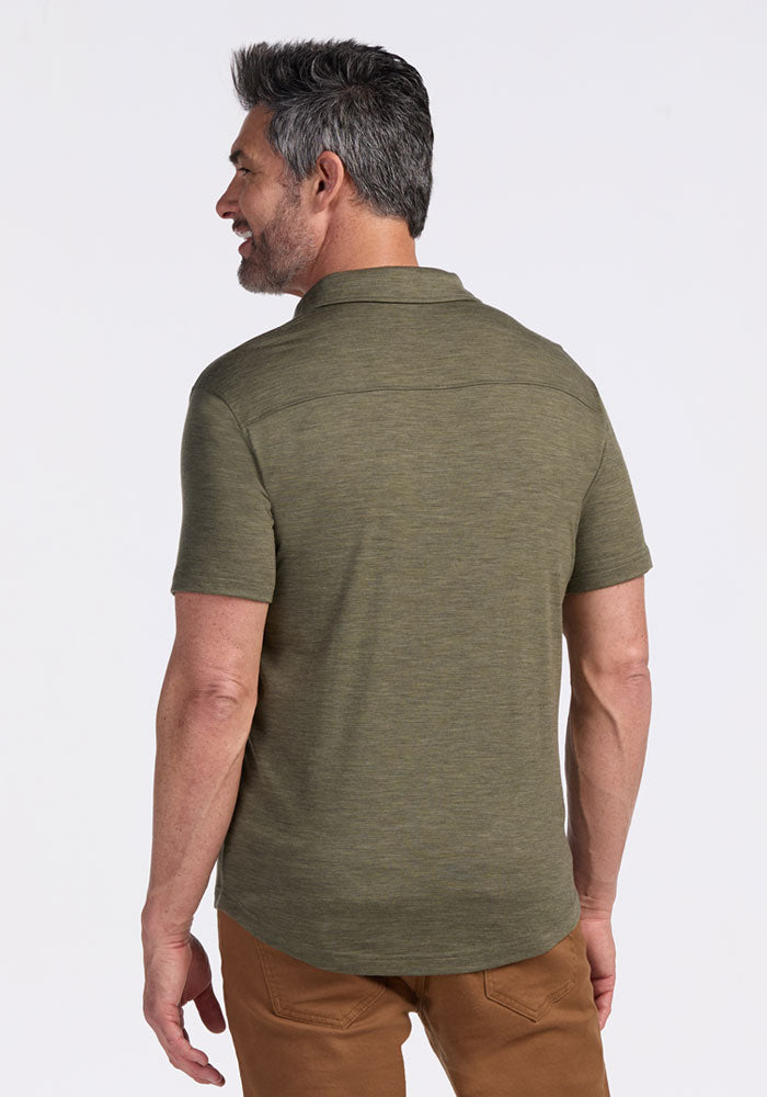 A man with gray hair and a beard, facing away from the camera, is wearing a Woolx Endi Full Button Down Shirt in Sage Olive Melange and brown pants against a plain white background.