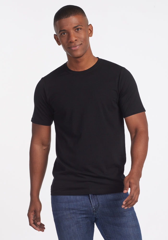 A man wearing a Woolx Endurance Tee in black and blue jeans stands smiling against a plain white background. | Trell is 6’2”, wearing a size M