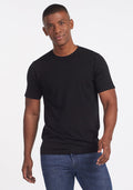 A man wearing a Woolx Endurance Tee in black and blue jeans stands smiling against a plain white background. | Trell is 6’2”, wearing a size M