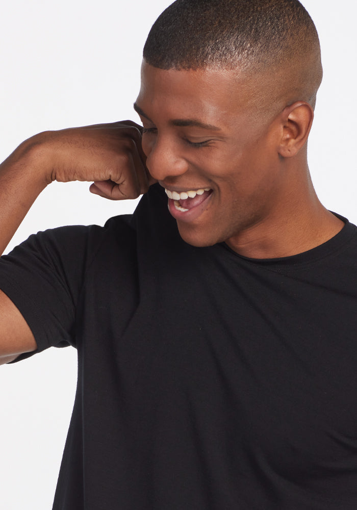 A person wearing a Woolx Endurance Tee - Black smiles while flexing their arm and looking down. The plain white background highlights the sleek design of this performance gear.