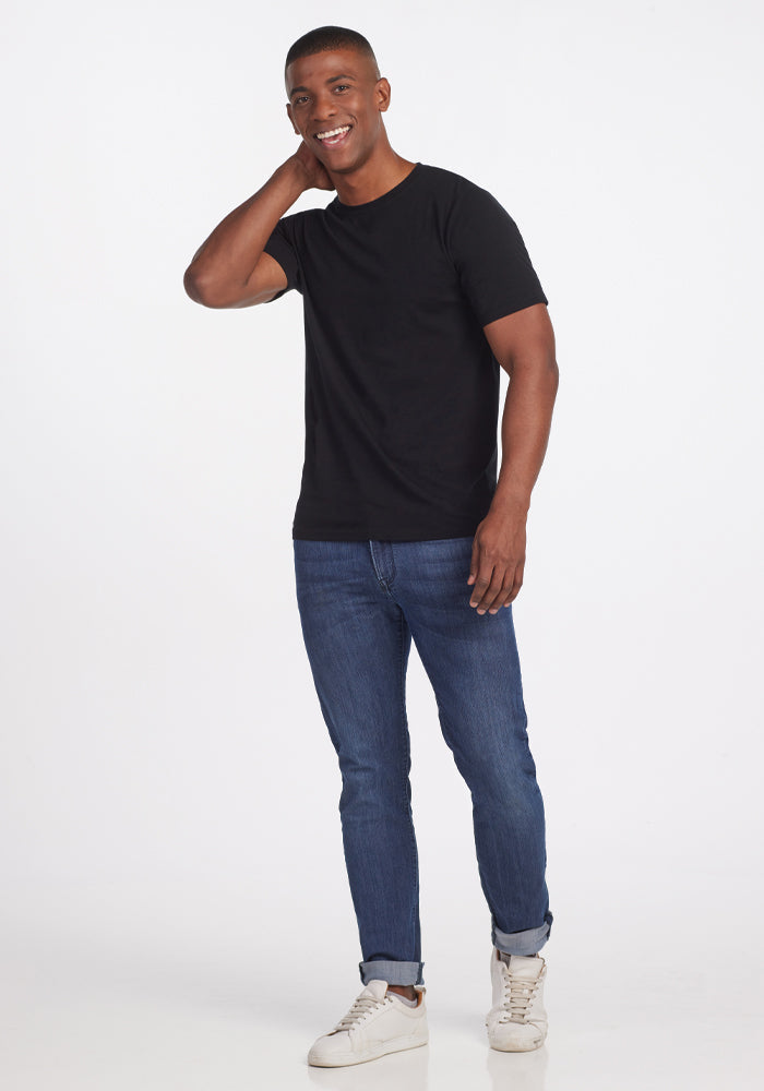 A person in a Woolx Endurance Tee - Black, blue jeans, and white sneakers stands smiling with one hand behind their neck against a plain white background.