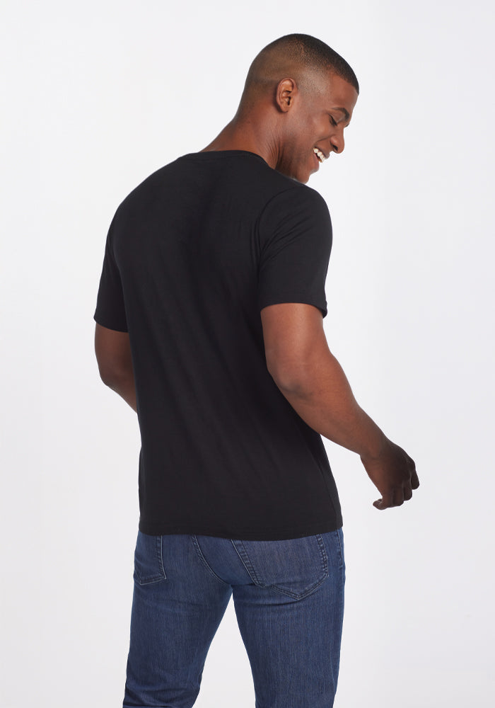 A person wearing a Woolx Endurance Tee in black and blue jeans is smiling and looking over their shoulder against a white background.
