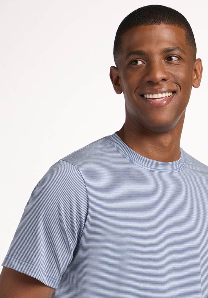 A young man with short hair smiles and looks to the side, wearing the Woolx Endurance Tee in Blue Sand Mini Stripe—ideal performance gear—posed against a plain, light background.