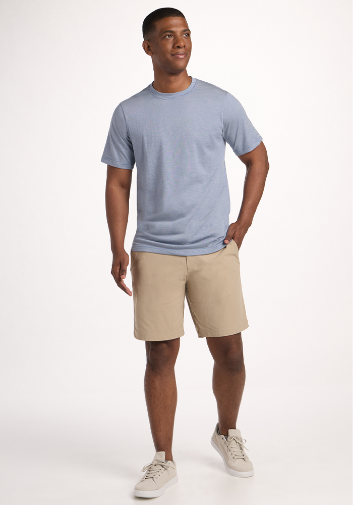 A man stands against a white background, wearing the Woolx Endurance Tee in Blue Sand Mini Stripe, beige shorts, and light gray sneakers. With one hand in his pocket, he gazes to the side with a relaxed look.