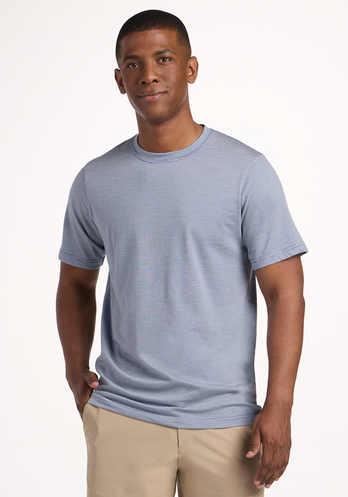 A man stands against a white background, wearing the Woolx Endurance Tee in Blue Sand Mini Stripe and beige pants. He smiles slightly with one hand in his pocket.