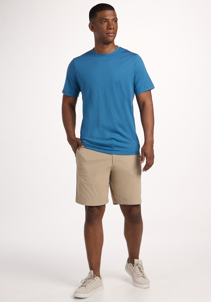 A man stands at an angle, wearing the Woolx Endurance Tee - Coastal Mini Stripe, beige shorts, and white sneakers. One hand is in his pocket as he looks off to the side against a plain white background.