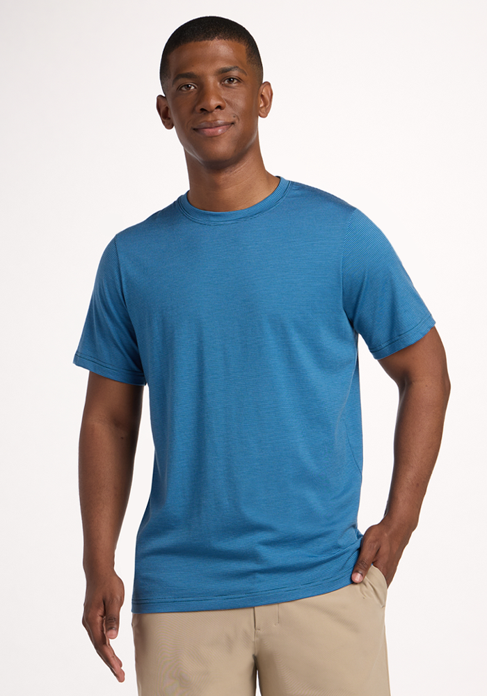 A smiling man stands against a plain background, wearing the Woolx Endurance Tee - Coastal Mini Stripe and light-colored pants, with one hand in his pocket—ideal versatile performance gear for any adventure.