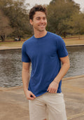 A smiling man in a Woolx Endurance Tee • Final Sale - Estate Blue and beige shorts stands by a pond on a cloudy day.
