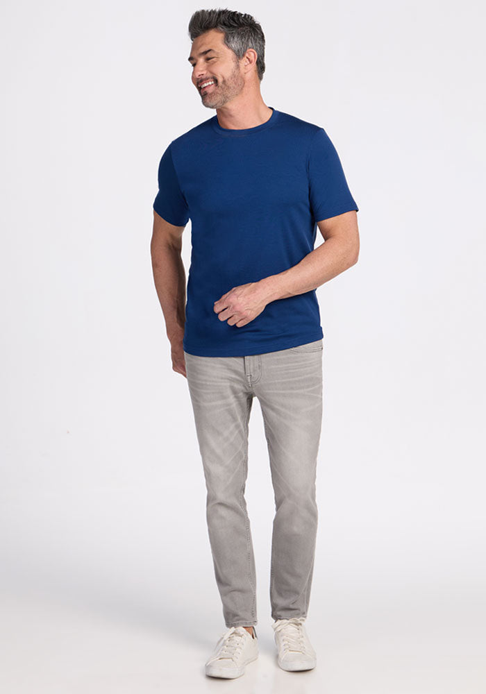Man in a Woolx Endurance Tee in Estate Blue, gray jeans, and white sneakers smiles against a plain white background.