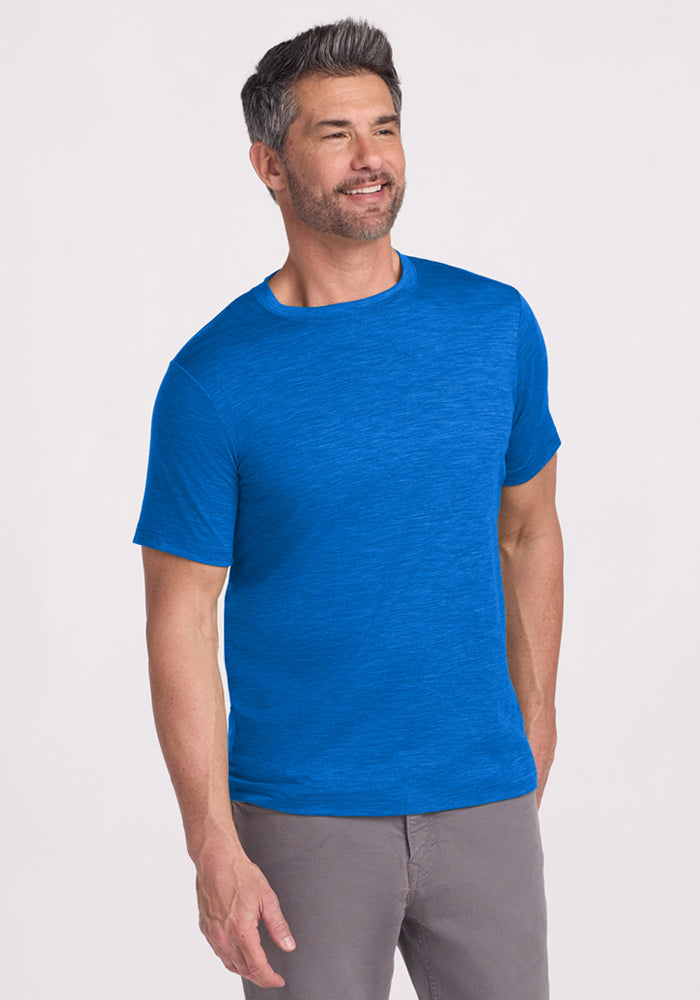 A smiling man wears the Woolx Endurance Tee in Glacial Blue and gray pants, standing against a plain light background.