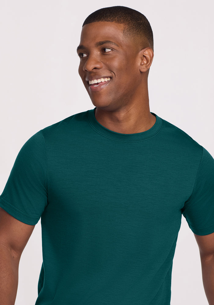 A smiling man wears a Woolx Endurance Tee in Riverbank Green, standing against a plain light background.