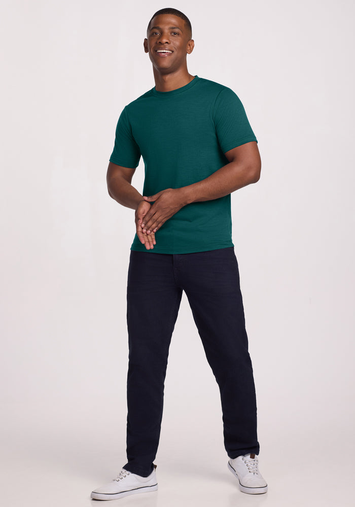 Man smiling in Woolx Endurance Tee in Riverbank Green, dark pants, and white sneakers against a plain background.