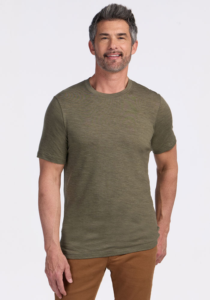 Smiling man in a Woolx Endurance Tee (Sage Olive Melange) and brown pants stands against a plain light background.