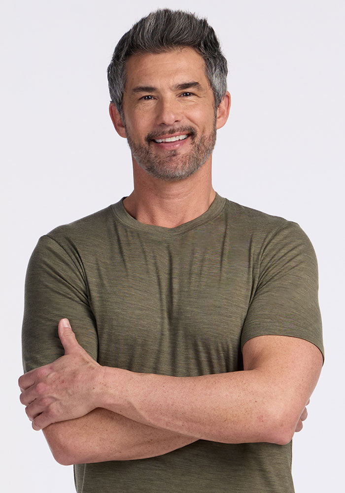 Smiling man with short gray hair wears a Woolx Endurance Tee in Sage Olive Melange, arms crossed, light background.