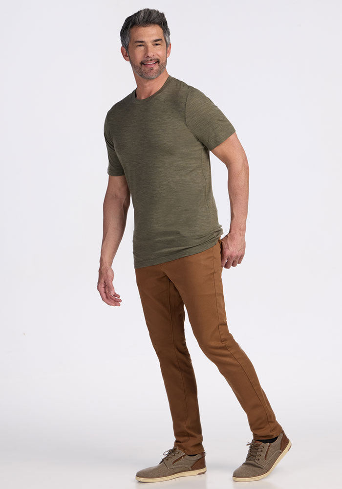 Man wearing a Woolx Endurance Tee in Sage Olive Melange, brown pants, and taupe sneakers, smiling against a white background.