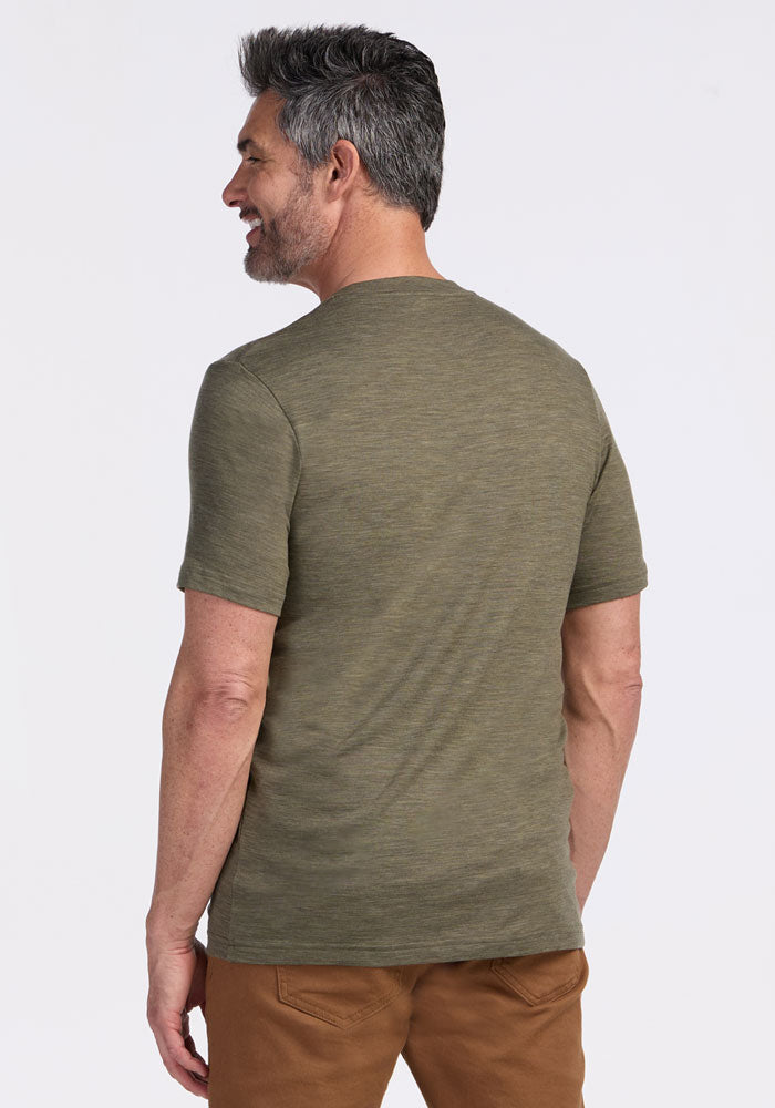 A man with gray hair, wearing a Woolx Endurance Tee in Sage Olive Melange and brown pants, stands smiling, facing away.