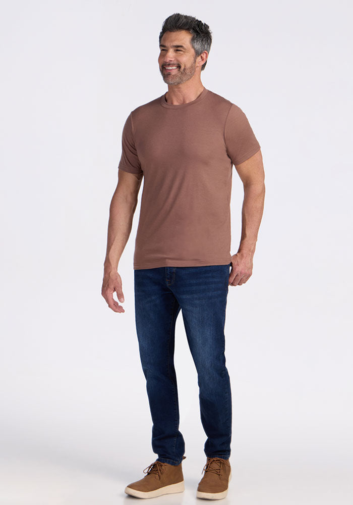 Man with short dark hair wears the Woolx Endurance Tee in Sienna Kiss, blue jeans, and brown shoes, smiling.
