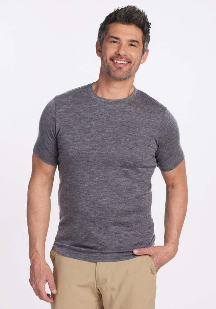 A person smiles while wearing a fitted Woolx Endurance Tee in Graphite Heather and beige pants, standing against a plain white background. They have short, dark hair with touches of gray and their right hand is in their pocket. | Matthew is 6’, wearing a size L