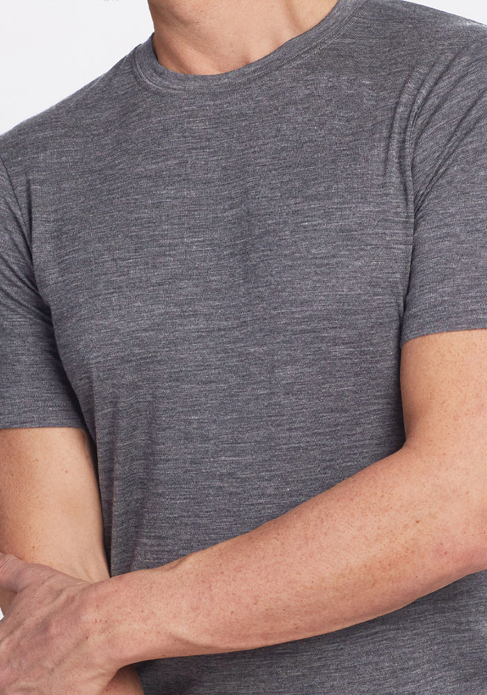 A person wearing a Woolx Endurance Tee in Graphite Heather stands with arms crossed against a plain background, highlighting the ideal fusion of comfort and style in performance gear.