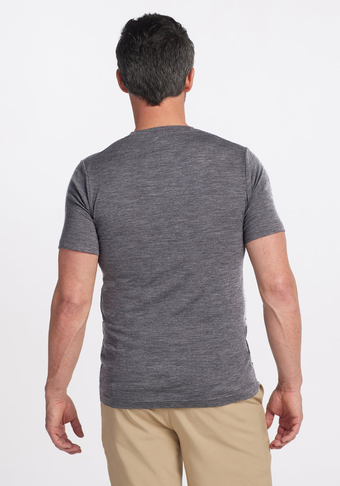 A man, clad in a Woolx Endurance Tee - Graphite Heather and khaki pants, stands with his back to the camera. The plain white background enhances the sleek design of his performance gear.