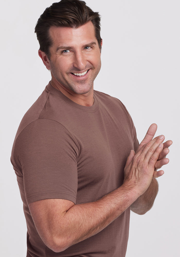 Smiling man in a Woolx Endurance Tee - Peppercorn stands with hands together, facing sideways against a plain background.