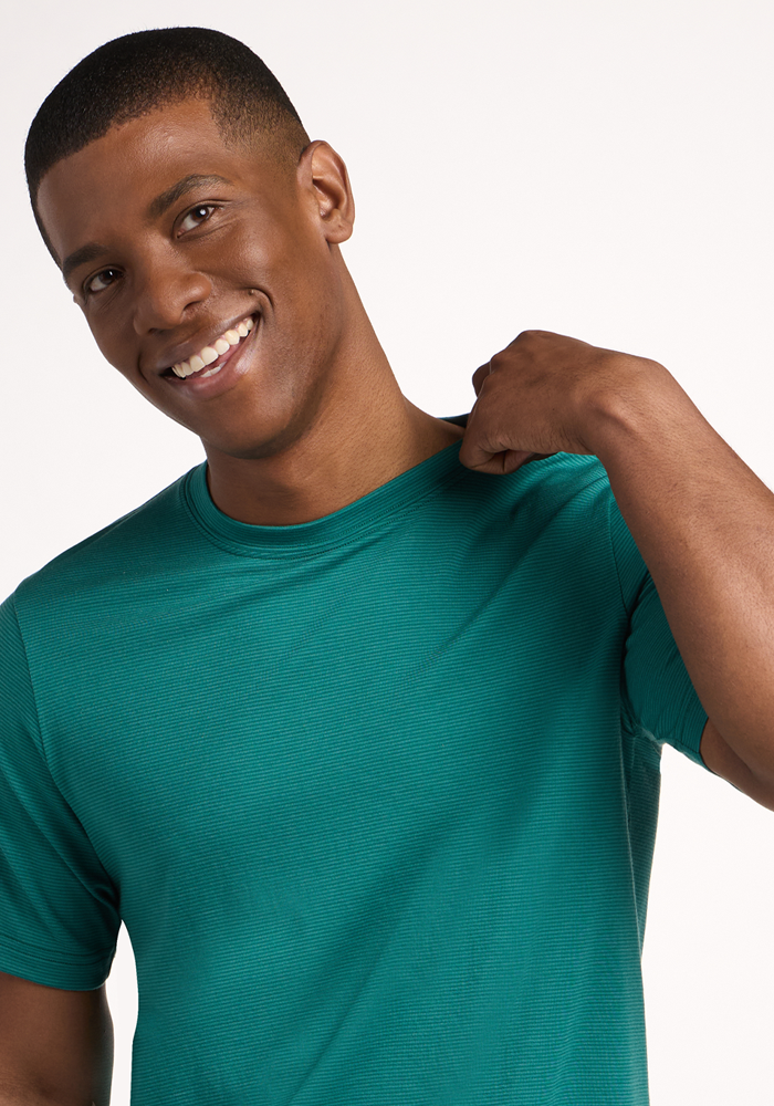 A smiling man in a Woolx Endurance Tee - Woodland Mini Stripe stands against a plain background, holding the back of his shirt collar with one hand.