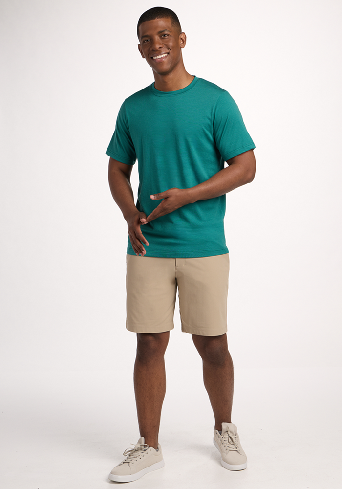 A man stands smiling against a plain white background, wearing the Woolx Endurance Tee - Woodland Mini Stripe, beige shorts, and light-colored sneakers. He gestures with his hands, looking relaxed and casual.