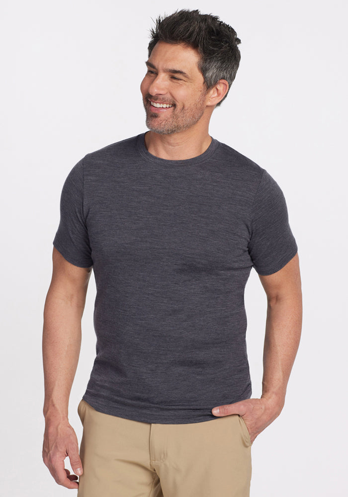 A man with short dark hair and light facial hair smiles while wearing the Woolx Endurance Tee in Cinder Melange and beige pants, standing against a plain white background. | Matthew is 6’, wearing a size L