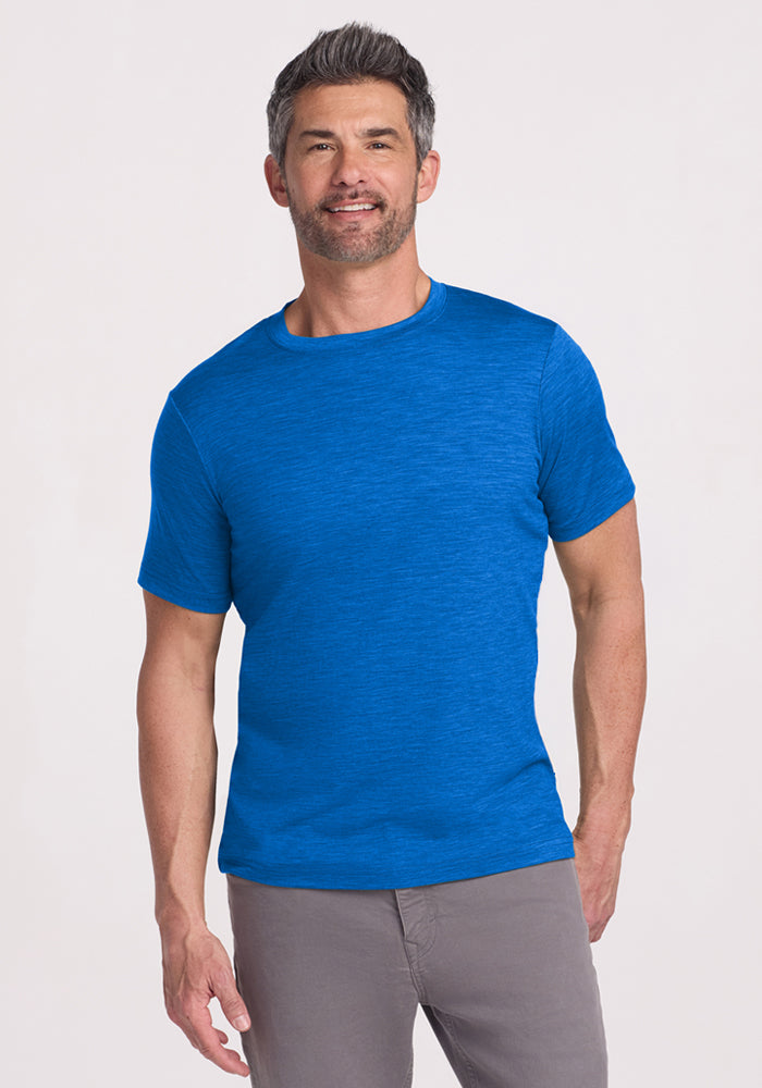 Smiling man wears Woolx Endurance Tee in Glacial Blue and gray pants, standing against a plain light background.