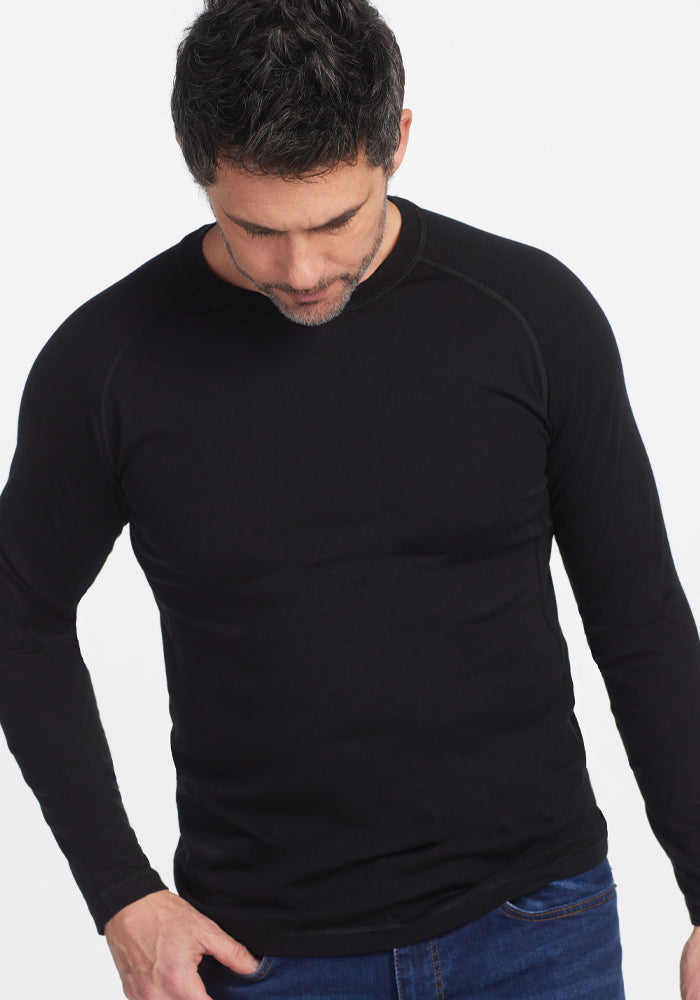 A man with short dark hair is looking down while wearing a black long sleeve Essential Tee from Woolx and blue jeans against a white background.