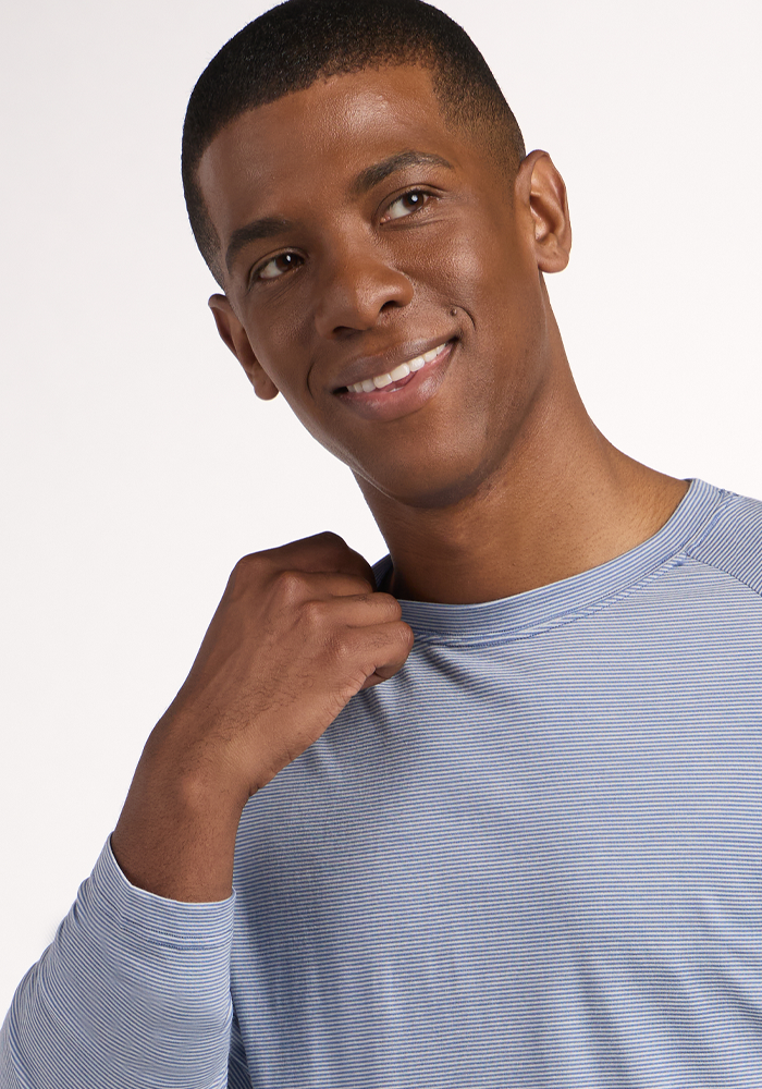 A man in the Woolx Essential Tee - Blue Sand Mini Stripe smiles and looks to the side, holding his shirt near his shoulder against a plain, light-colored background.