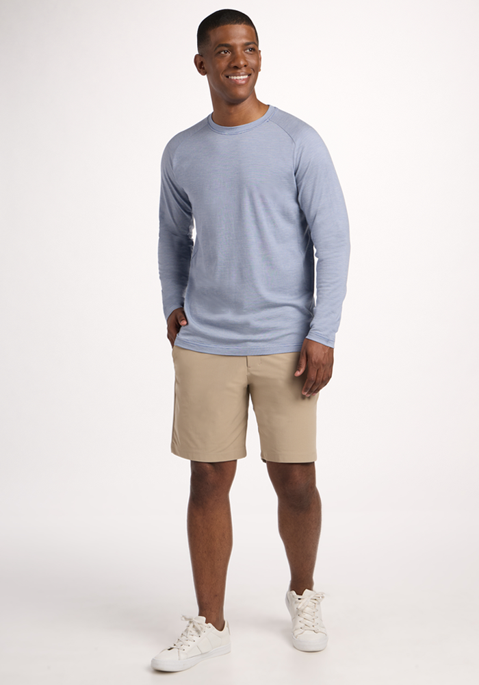 A man smiles, wearing the Woolx Essential Tee in Blue Sand Mini Stripe, beige shorts, and white sneakers. The temperature-regulating Merino wool fabric keeps him comfortable against a plain white background.