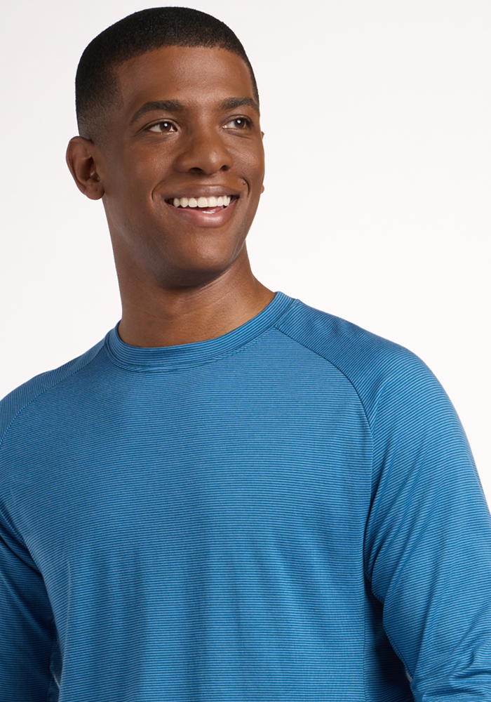 A smiling man with short hair wears the Woolx Essential Tee - Coastal Mini Stripe, a blue Merino wool long sleeve crew-neck shirt, and looks slightly to the side against a plain light background.