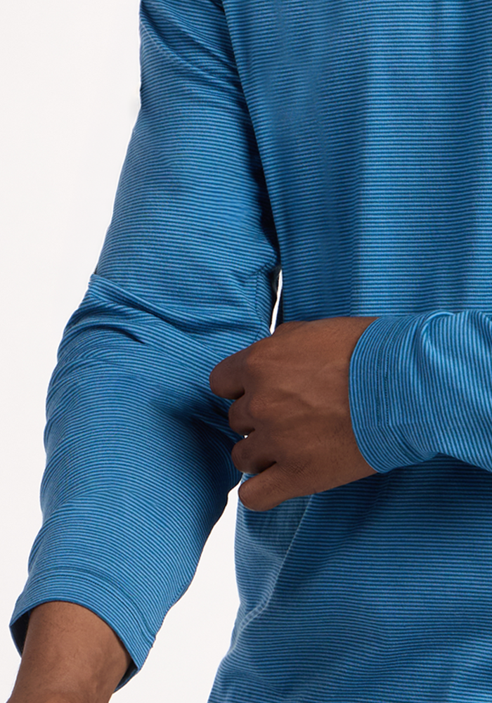 A person wearing a blue Woolx Essential Tee - Coastal Mini Stripe adjusts the sleeve on their left arm, with a plain, light-colored background.