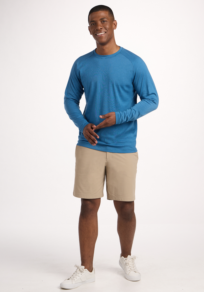 A smiling man with short hair stands against a plain background, wearing the Woolx Essential Tee in Coastal Mini Stripe, tan shorts, and white sneakers. He holds his hands together in front of him.