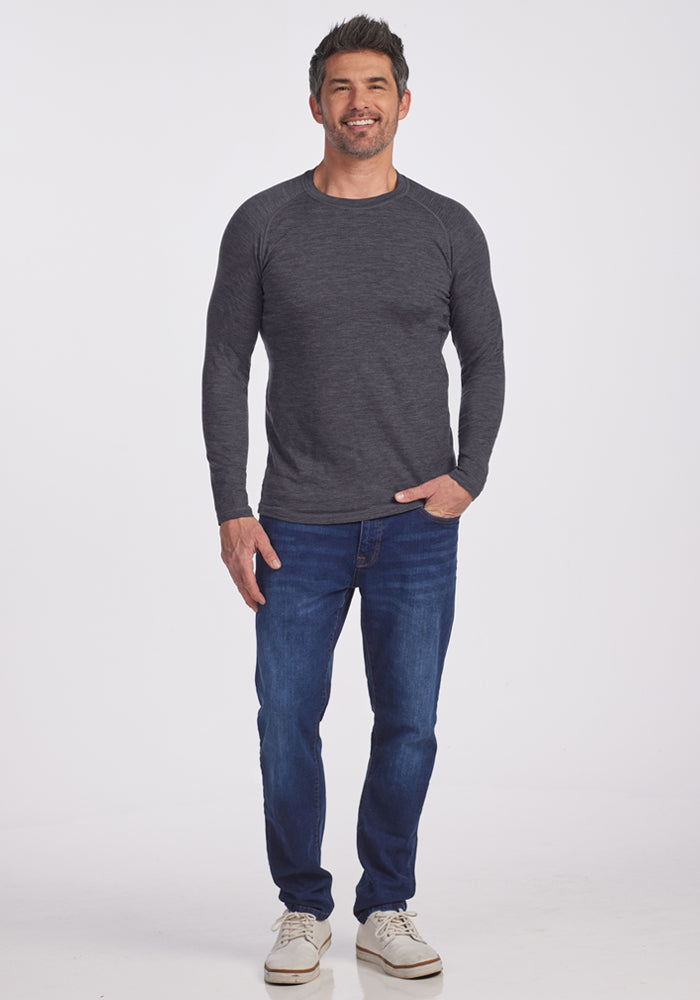 A man in a Woolx Essential Tee in Cinder Melange, blue jeans, and white sneakers smiles against a white background.