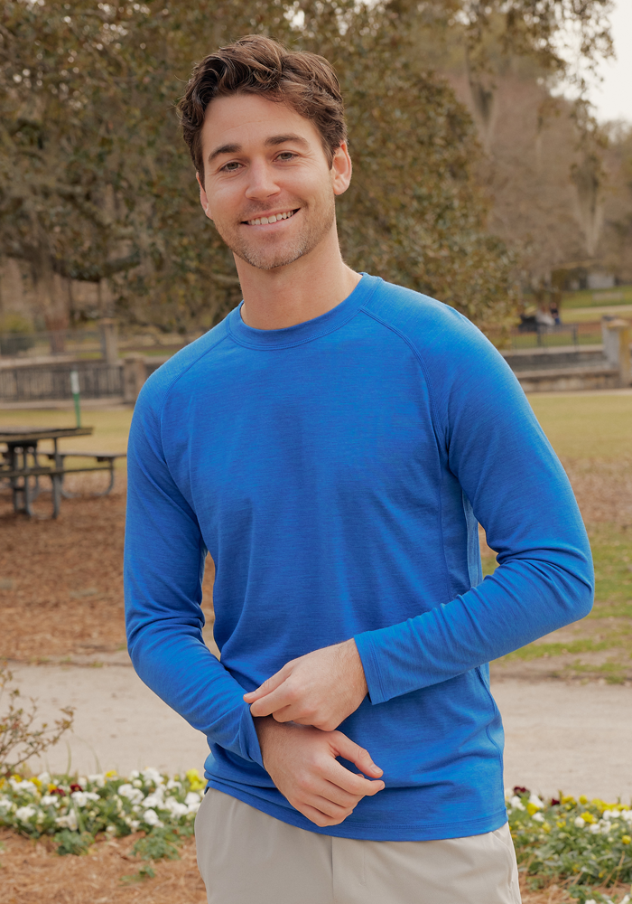 A man in the Woolx Essential Tee • Final Sale - Glacial Blue smiles while outdoors in a park with trees and flowers.