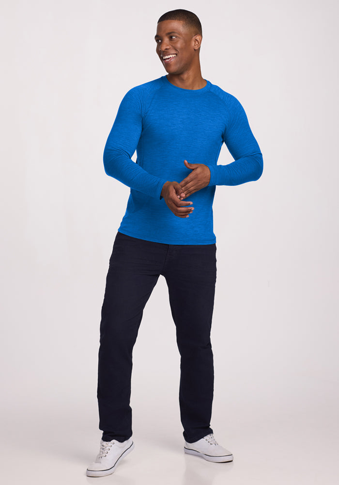 Man in a Woolx Essential Tee in Glacial Blue, dark pants, and white sneakers standing and smiling against a plain background.