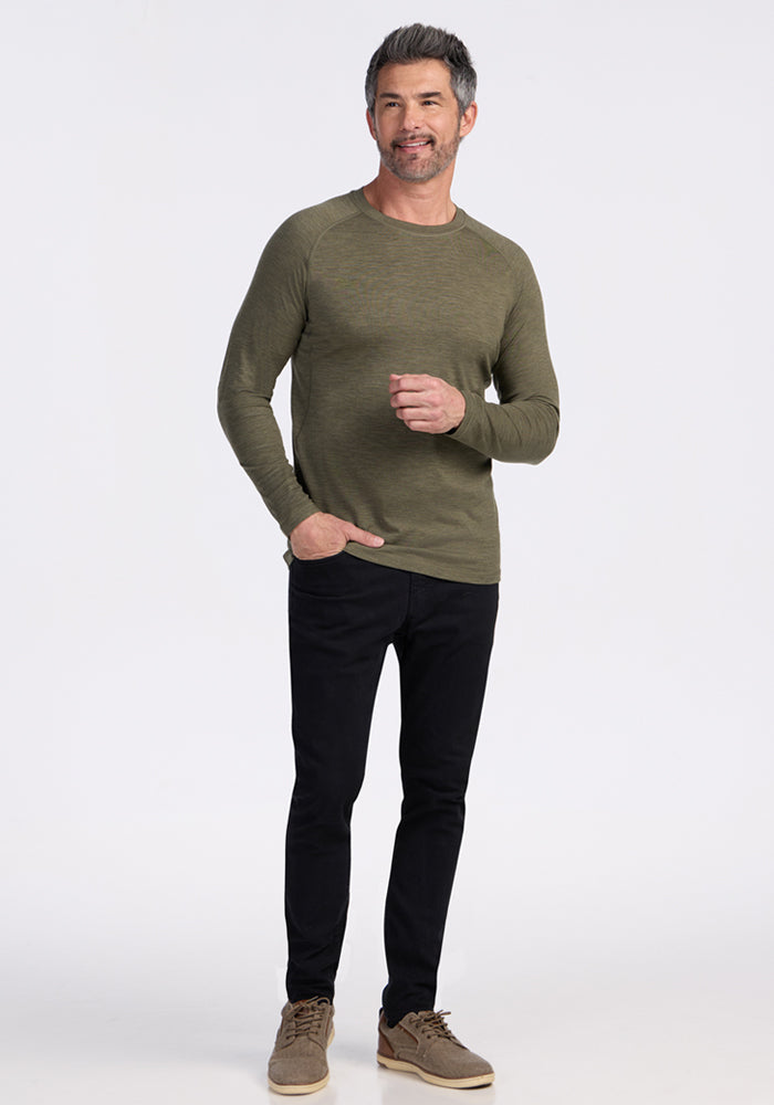 Man in a Woolx Essential Tee in Sage Olive Melange, black jeans, and brown shoes smiles against a white background.