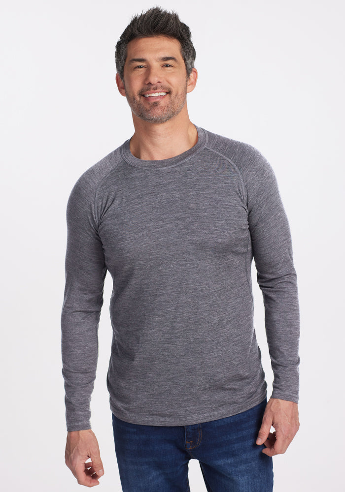 A man with short dark hair and stubble smiles while wearing a Woolx Essential Tee in Graphite Heather and blue jeans, posing against a plain white background. | Matthew is 6’, wearing a size L