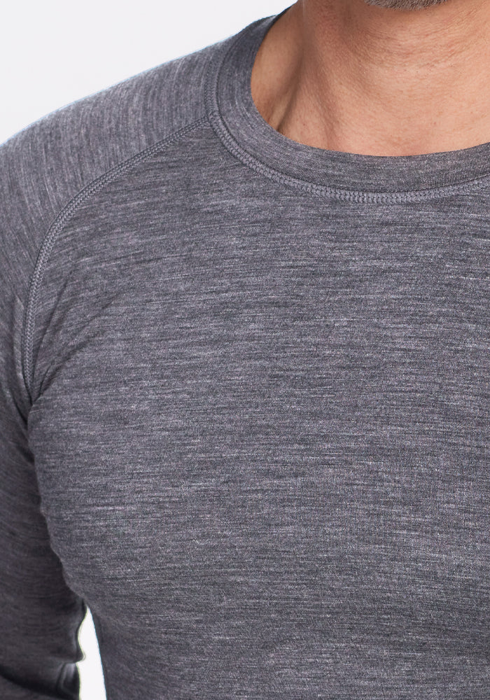 A close-up shows a person wearing the Woolx Essential Tee in Graphite Heather, revealing its textured appearance. Only their upper torso and part of the neck are visible against a white background, emphasizing its temperature-regulating properties.