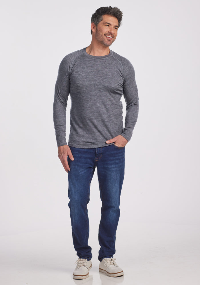 A man with short hair smiles on a plain white background, wearing the Woolx Essential Tee in Graphite Heather instead of a long-sleeve gray shirt and blue jeans. One hand is in his pocket, and he has white sneakers on his feet.