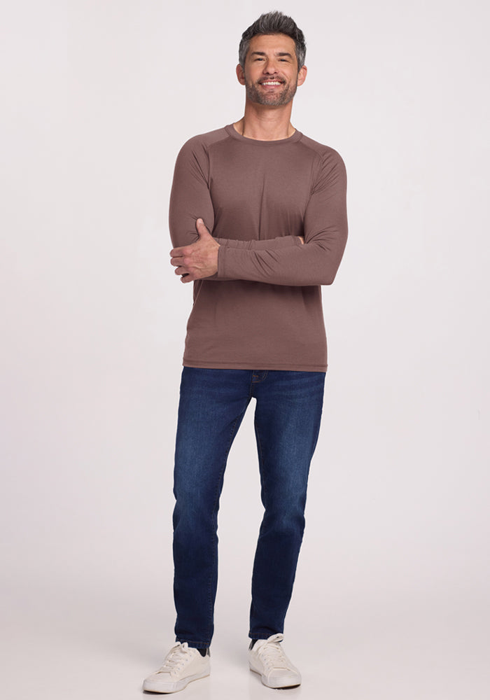 A man in a Woolx Essential Tee - Peppercorn, blue jeans, and white sneakers smiles against a plain background.